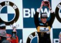 Ayrton Senna, the legendary racing driver, triumphantly holds a trophy above his head on a podium adorned with BMW logos. Wearing a Nacional and Goodyear-branded suit, he stands alongside another victorious driver, celebrating their Lotus F1 victory with matching trophies in hand.