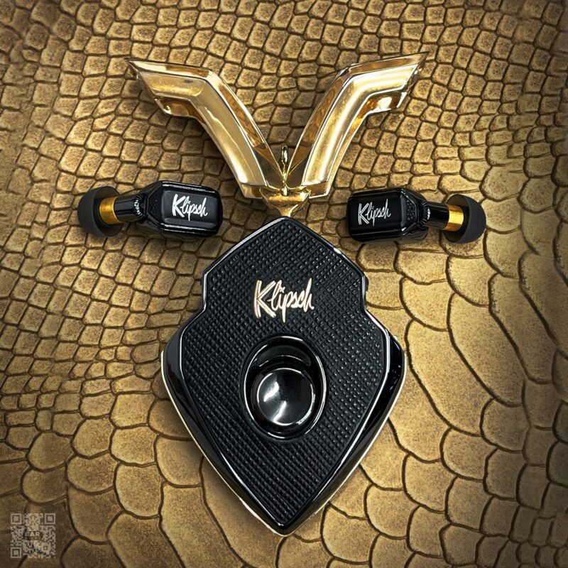 Black and gold Klipsch earbuds and case arranged on a textured, gold, reptile-patterned surface with a gold V-shaped ornament above them—perfect luxury accessories to upgrade your style.