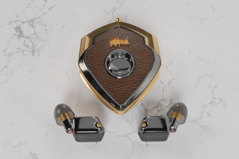 A pair of black and gold wireless earbuds with a shield-shaped, brown and gold charging case on a light marble surface—perfect for a style upgrade in luxury accessories.