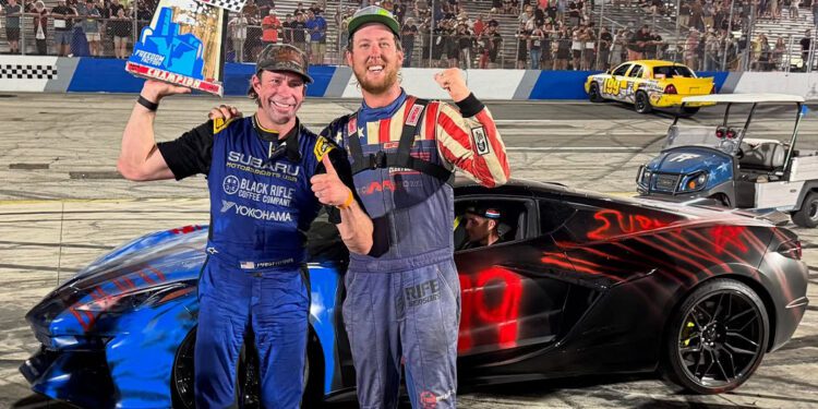 Two race car drivers stand smiling on the racetrack. One holds a trophy in front of a C8 Corvette Z06, its sleek black and blue design gleaming. Excited crowds cheer from the stands, still buzzing with energy after an unforgettable Freedom 500 Race.
