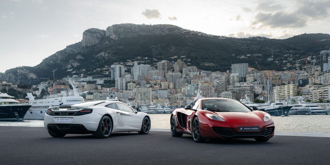 Two sports cars, one white and one red, are parked on a waterfront with yachts and a cityscape backed by hills—evoking the excitement of a McLaren auction and the racing spirit of Lewis Hamilton.