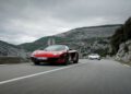 Two sports cars, one red and one white, speed along a winding mountain road under cloudy skies—reminiscent of McLaren legends like Jenson Button and Lewis Hamilton pushing limits on challenging terrain.
