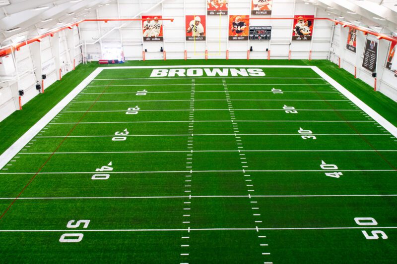 Indoor football practice field with artificial turf, white yard lines, and "BROWNS" written in the end zone; sports banners visible on the far wall.