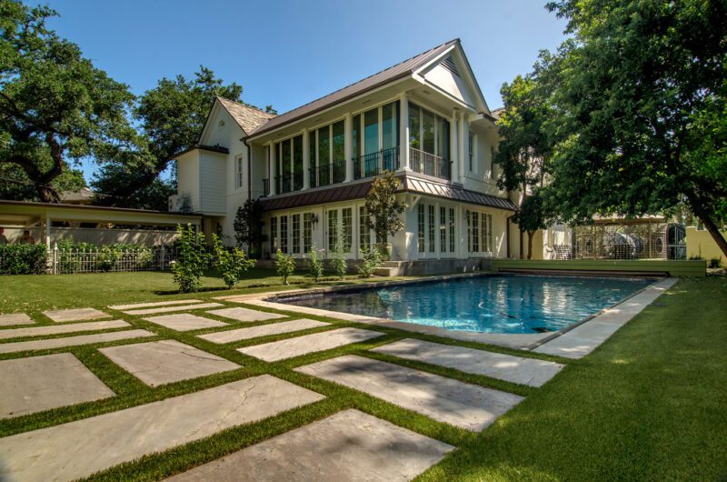 A modern two-story house with large windows overlooks a rectangular swimming pool, surrounded by a manicured lawn and stone pavers.
