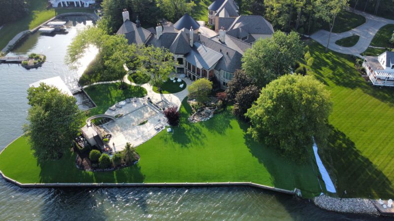 Aerial view of a large waterfront house with landscaped yard, swimming pool, dock, and surrounding trees on a sunny day.
