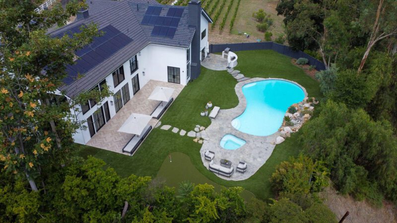 Aerial view of a modern house with solar panels, a swimming pool, hot tub, patio area, putting green, and landscaped yard surrounded by trees and vineyards.