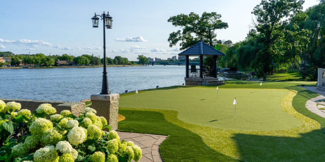 A landscaped backyard features a putting green, a gazebo near the water, flowering hydrangea bushes, and a lamppost under a clear sky.