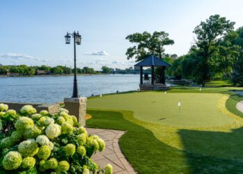A landscaped backyard features a putting green, a gazebo near the water, flowering hydrangea bushes, and a lamppost under a clear sky.