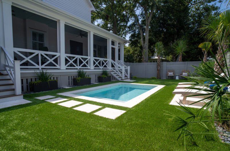 A small backyard pool with white stone pavers surrounded by green artificial grass, adjacent to a white house with a screened porch and outdoor seating area.