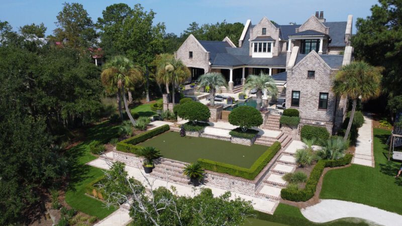 Large brick estate with multiple gables, landscaped gardens, manicured hedges, and palm trees, viewed from above on a sunny day.