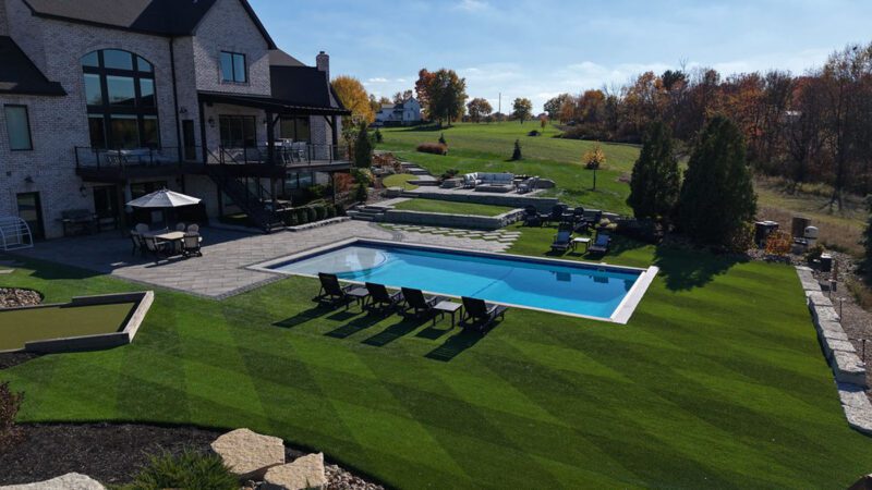 A large backyard features a rectangular swimming pool, patio seating areas, lounge chairs, and manicured green lawns behind a two-story brick house.