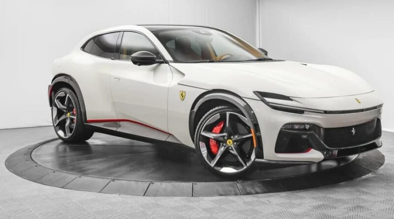 A white Ferrari Purosangue SUV is displayed in a showroom, featuring black and red accents, large black alloy wheels, and red brake calipers. Discover this stunning Ferrari Purosangue for sale.