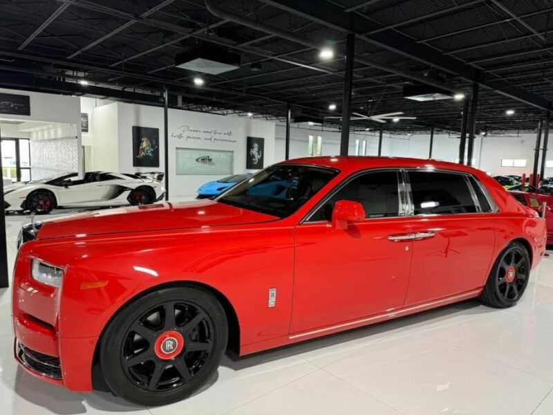 A red Rolls-Royce sedan with black wheels is parked indoors alongside other luxury cars in a modern showroom, making it a standout exotic car for sale.