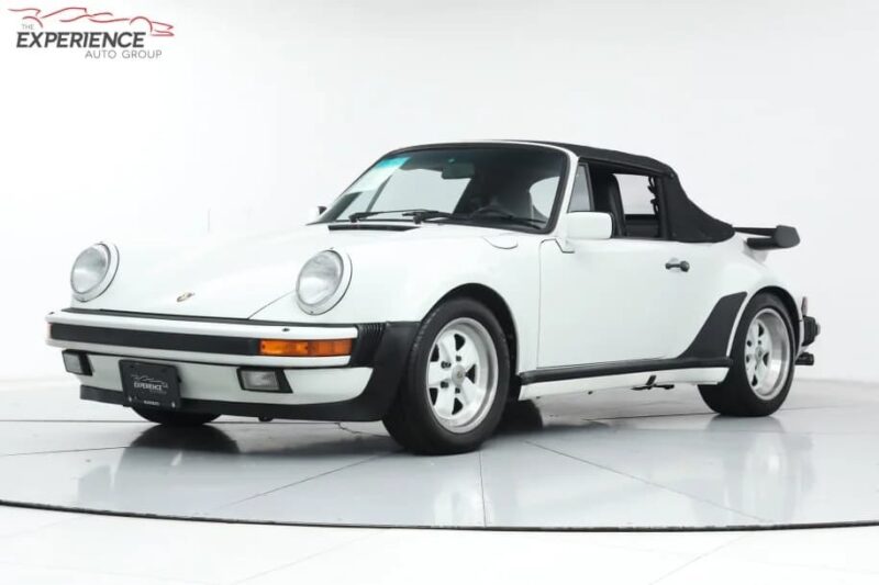 A white classic Porsche 911 convertible with black soft top, viewed from the front left, parked in a studio setting—an iconic luxury car for sale that exudes timeless elegance.