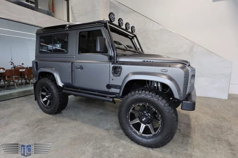 A matte gray Land Rover Defender, a true luxury car with large off-road tires and roof-mounted lights, is parked indoors on a concrete floor.