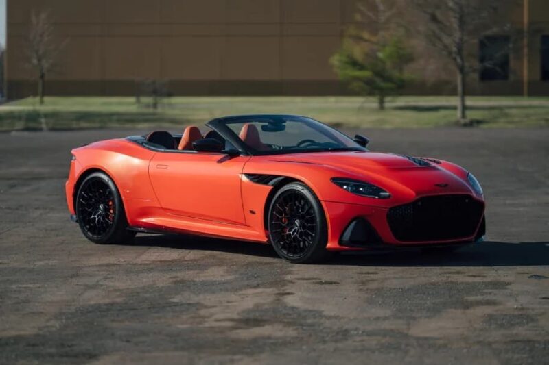 A bright orange Aston Martin DBS 770 Ultimate grand touring convertible sports car is parked on an empty asphalt surface with a blurred building and trees in the background.
