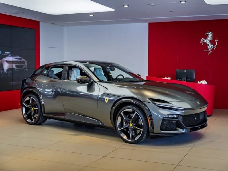 A gray Ferrari Purosangue SUV is displayed in a showroom with red and white walls, Ferrari logos, and a digital screen in the background.