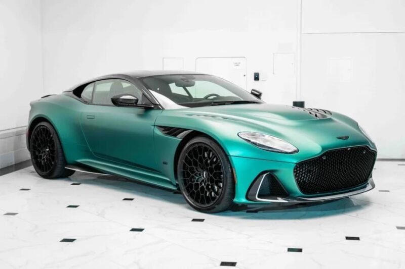 A green Aston Martin DBS 770 Ultimate sports car is parked indoors on a white marble floor with black accents, set against a plain white wall—an icon of Grand Touring elegance.