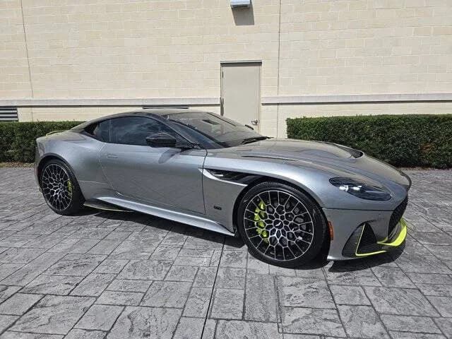 A silver Aston Martin DBS with black and yellow-accented wheels is parked on a stone driveway in front of a light-colored wall and lush greenery, embodying the essence of grand touring sophistication.
