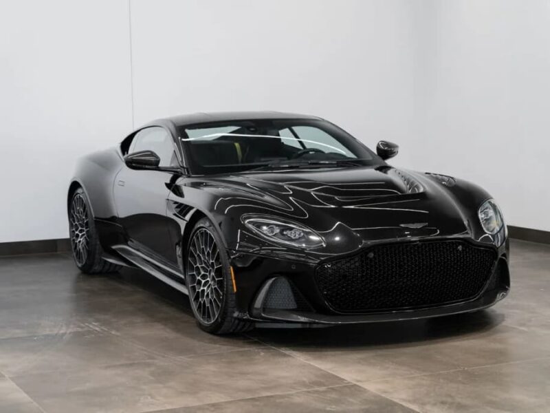 A black Aston Martin DBS grand touring sports car is parked indoors on a polished concrete floor, positioned at an angle facing left.