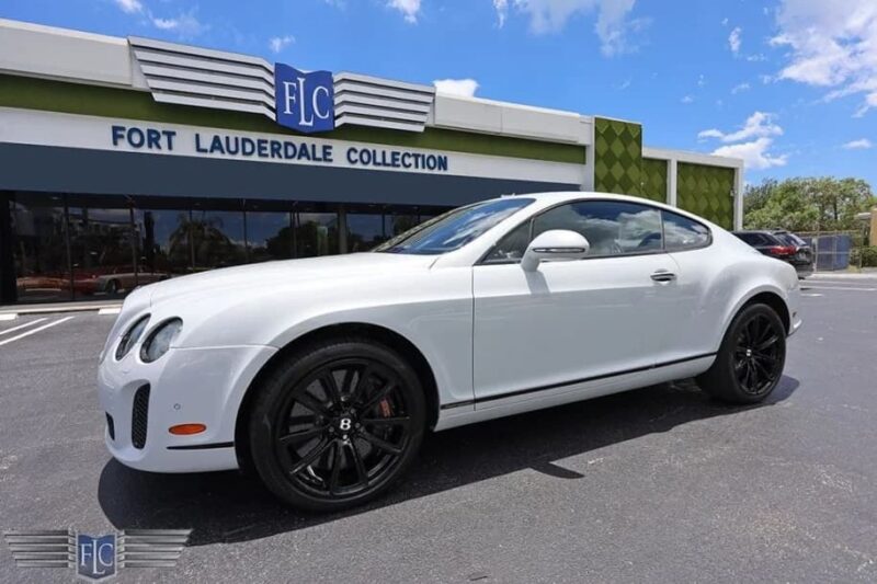 A white Bentley coupe with black wheels, a true luxury car, is parked outside Fort Lauderdale Collection under a clear blue sky.