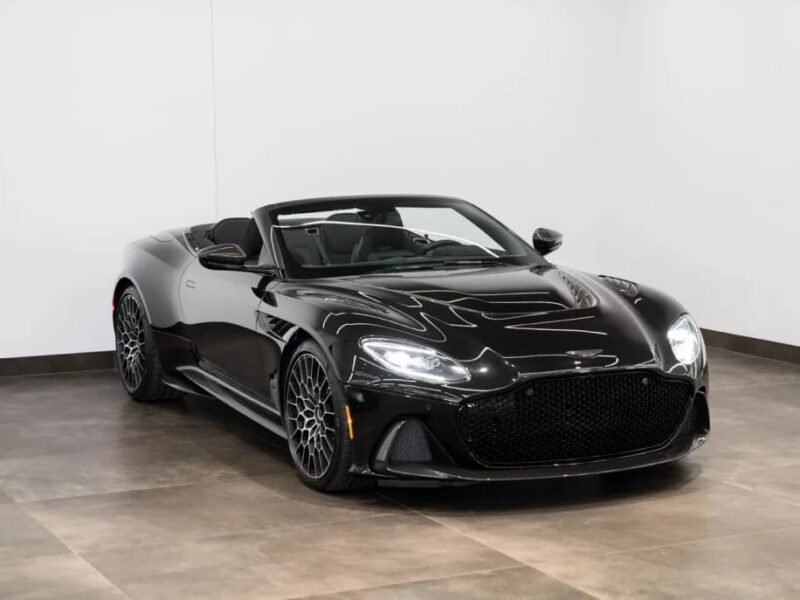 A black Aston Martin DBS grand touring convertible with the top down is parked indoors on a tiled floor against a plain white wall.