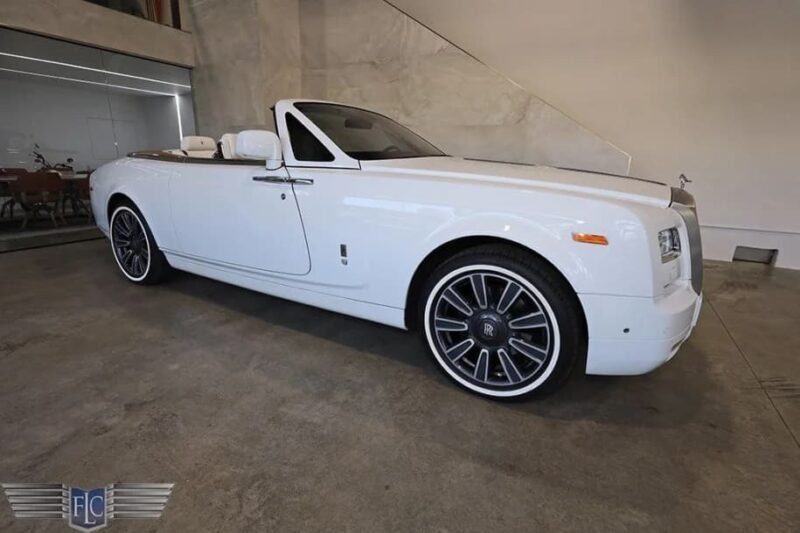 A white Rolls-Royce convertible, a true luxury car for sale, is parked indoors on a concrete floor and viewed from the side at a slight angle.