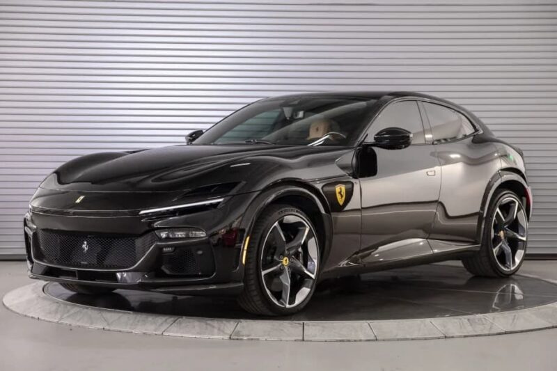 A black Ferrari Purosangue SUV is parked indoors on a turntable, with a silver corrugated metal wall in the background—an impressive Ferrari SUV ready for those seeking a Ferrari Purosangue for sale.