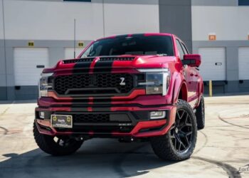 A red Ford F-150 pickup truck with black racing stripes is parked in front of a modern industrial building with white garage doors. Shelby Trucks enthusiasts take note—this standout vehicle is for sale.