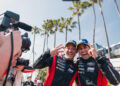 Two race car drivers in red jackets celebrate a thrilling one-two finish, holding three fingers up, surrounded by cameras and palm trees in Long Beach.