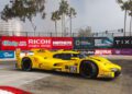 A yellow Porsche race car with number 85 speeds through the Long Beach street circuit, surrounded by sponsor banners and palm trees in the background, as it races towards the finish.