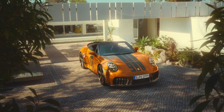 A limited edition orange Porsche 911 convertible with black racing stripes is parked on a paved driveway in front of a modern white house, surrounded by greenery.