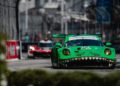 A sleek green Porsche with a fierce teeth design on the front speeds along the Long Beach racetrack, leading towards a potential one-two finish as a red car follows closely. Blurred spectators eagerly watch from behind a chain-link fence.
