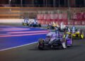 Race cars, including a sleek Porsche, speed on the curved Long Beach track with barriers lining the sides under bright artificial lights. Multiple vehicles follow closely behind, battling for a thrilling one-two finish.