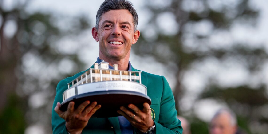 A person in a green jacket holds a trophy shaped like a building, reminiscent of Rory McIlroy's Masters win, standing outdoors with trees in the background.