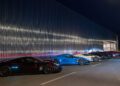 A row of seven parked sports cars in various colors, including black, blue, white, and red, are aligned beside a modern, metallic building at night.