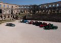 A line of Rimac sports cars, their open doors like wings, forms a semicircle inside the ancient amphitheater. The Nevera stands apart on the left, a solitary marvel in the Trailblazers Tour.
