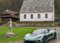 A sleek, modern Rimac sports car is parked on a paved driveway in front of an old-fashioned white building with a thatched roof, near a statue and surrounded by trees and grass.