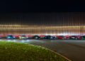 A lineup of ten sports cars, including Rimac Neveras, glows under the night sky as part of the Trailblazers Tour. Their headlights illuminate the ribbed metallic wall behind them, casting a mesmerizing glow over the grassy foreground.