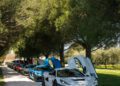 A row of Rimac Nevera sports cars with open butterfly doors glisten under the sun, parked on a tree-lined gravel road, as part of the exclusive Trailblazers Tour.