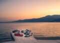 A group of sports cars, including the striking Rimac Nevera, is parked on a pier during sunset, overlooking a calm body of water with mountains in the background—perfect setting for a Trailblazers Tour.
