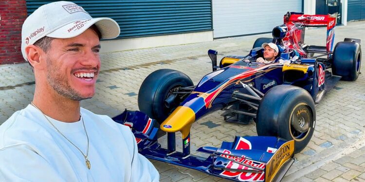 A man in a white shirt and cap grins beside a Red Bull F1 car, dreaming of the Bugatti purchase he'll enjoy on the autobahn.