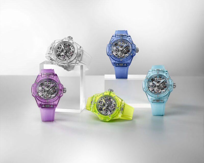 In celebration of Hublot's 20th Anniversary, five colorful, transparent wristwatches are displayed on a white surface. Each Big Bang watch showcases a visible intricate mechanism, with bands in shades of purple, blue, yellow, and white.