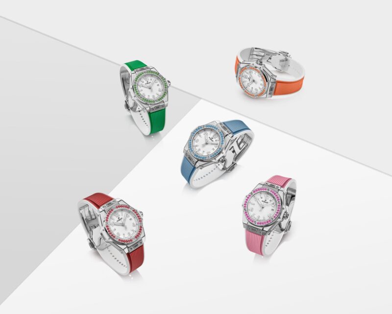 Five wristwatches with white faces, silver bezels, and colorful bands in green, orange, blue, red, and pink are arranged on a reflective surface. In celebration of Hublot’s 20th anniversary, this collection pays tribute to the vibrant spirit reminiscent of the Big Bang.
