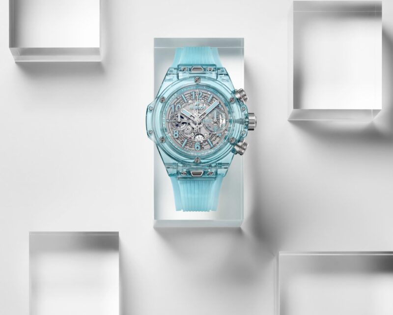 Displayed on a white geometric background is a Hublot wristwatch, featuring a transparent light blue design with visible mechanical movement. This timepiece evokes the spirit of the Big Bang collection, adding an iconic touch to anniversary watches.
