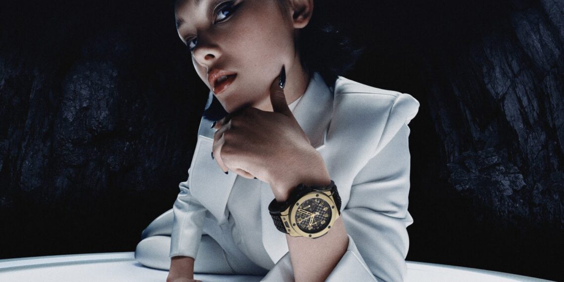 A person in a white suit poses dramatically, resting their chin on one hand, highlighting a large Hublot Big Bang wristwatch. The golden timepiece marks the 20th Anniversary of style against a dark, textured background.