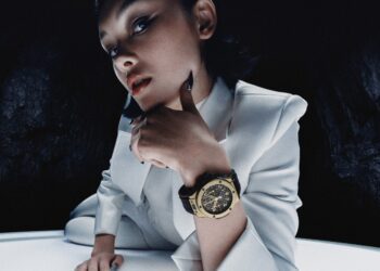 A person in a white suit poses dramatically, resting their chin on one hand, highlighting a large Hublot Big Bang wristwatch. The golden timepiece marks the 20th Anniversary of style against a dark, textured background.