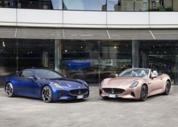 A blue Maserati GranTurismo coupe and a pink convertible sports car are parked side by side in front of a modern glass building in Modena.
