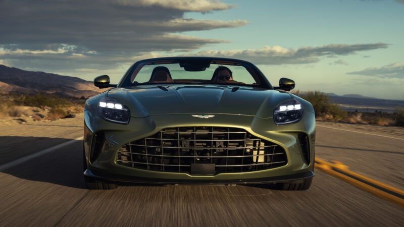 A green convertible sports car with its headlights on drives on a desert road under a partly cloudy sky.