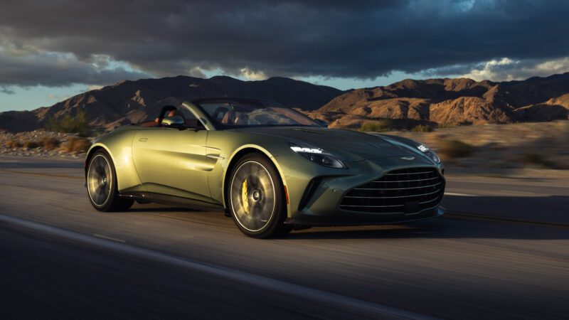 A green convertible sports car drives on a desert road with mountains and a cloudy sky in the background.